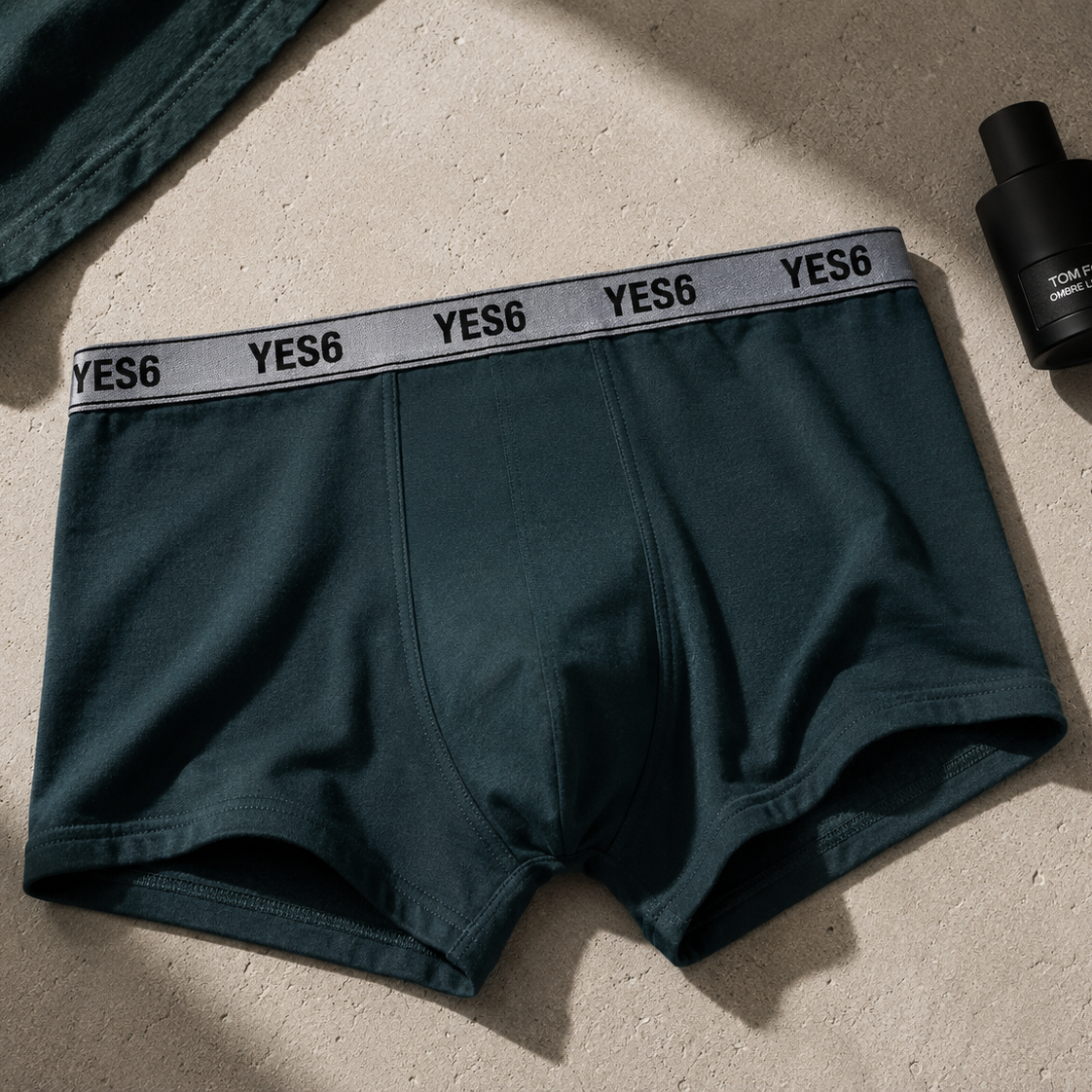 Dark teal green YES6 men's boxer briefs with a gray elastic waistband featuring the black YES6 logo, laid flat on a textured beige surface next to a black bottle.