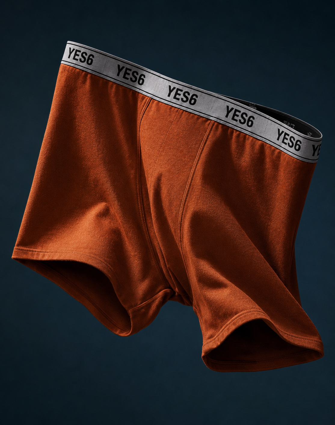 A pair of men's boxer briefs in rust orange, featuring a wide gray elastic waistband with the brand name 'YES6' printed repeatedly in black. The underwear is shown floating against a dark navy blue background.