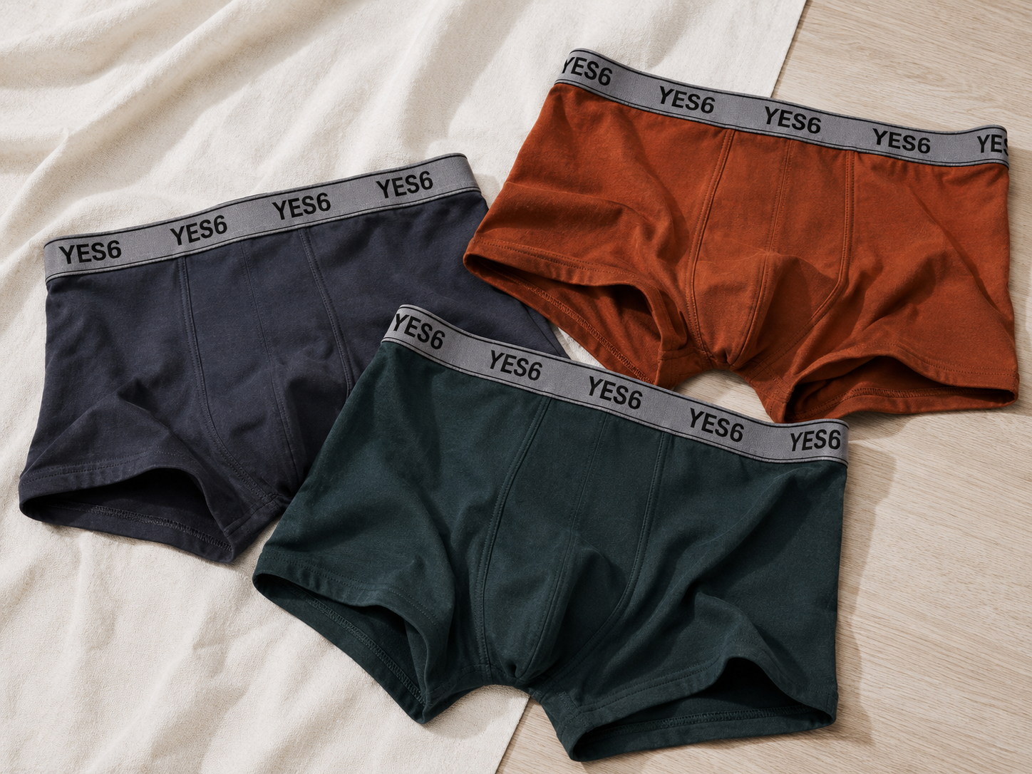 Three pairs of men's briefs laid flat on a light fabric surface: one dark navy, one rust orange, and one dark green. Each pair has a gray elastic waistband with the brand name YES6 repeated in black.